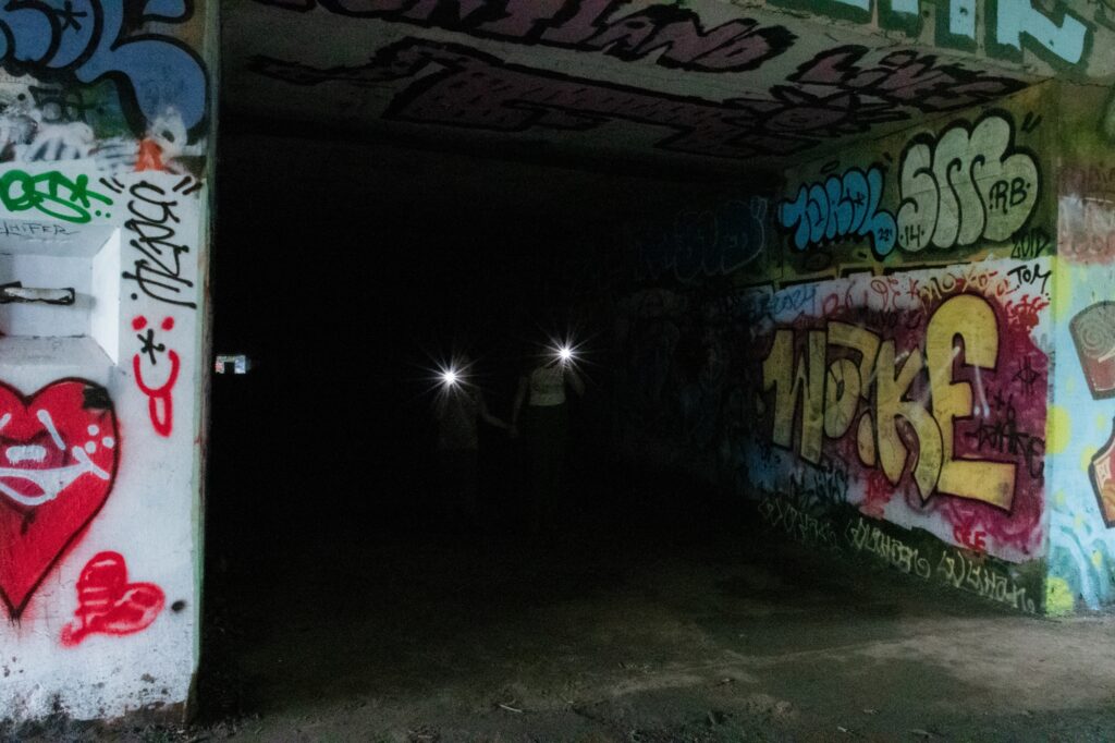Two people carrying lights emerge from a dark tunnel that is lined with graffiti